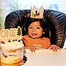 baby, child, birthday, cake, crown, high_chair, smiling, portrait, indoors, celebration, first_birthday, orange_shirt, dessert, frosting, fruit, party, happy, seated, backdrop, photography