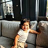 Emiliano is registered to the contest to win money with this photo: toddler, child, smiling, sitting, sofa, couch, living_room, indoor, portrait, sunlight, window, modern_interior, furniture, white_shirt, shorts, curly_hair, happy, reaching, kneeling, family_photo