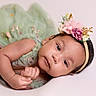 baby, child, infant, portrait, lying_down, flower_crown, headband, floral_outfit, green_dress, pink_background, studio_photo, close_up, face, eyes, smiling, hands, soft_lighting, pastel_colors, newborn_photography, adorable