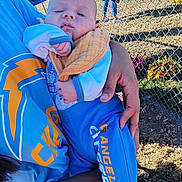Wrangler is registered to the contest to win money with this photo: baby, child, person, hand, clothing, sportswear, blue, orange, field, grass, fence, chain_link_fence, outdoor, daytime, adult, holding, team_logo, los_angeles_chargers, casual, relaxed