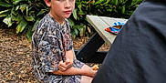 Chevy is registered to the contest to win money with this photo: child, boy, outdoor, picnic_table, bench, snack, camouflage_clothing, plants, greenery, leaves, wood_chips, person, eating, casual, daylight, nature, relaxed, seated, short_hair, expression