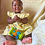 baby, child, yellow_dress, toy, colorful_toy, smile, happy, clapping_hands, barefoot, blanket, heart_pattern, sitting, cute, indoors, portrait, infant, soft_lighting, playful, comfort, bunny_prints