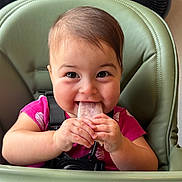Eleanor is registered to the contest to win money with this photo: baby, child, high_chair, popsicle, smiling, happy, indoor, face, hands, food, cute, young_child, seat, clothing, pink_shirt, skin, hair, table, infant, playful