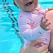 Sienna is registered to the contest to win money with this photo: baby, child, smiling, swimsuit, flamingo_pattern, water, pool, adult_hand, happy, wet_hair, skin, outdoor, summer, playful, splash, cute, person, holding, young_child, fun