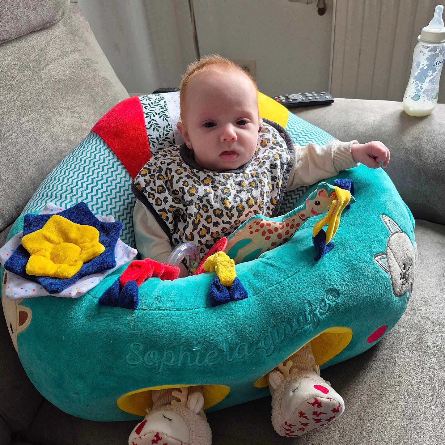 Noah a rejoint le concours — aidez-le/la à gagner de superbes lots ! baby, baby_bottle, bib, child, comfortable, couch, cozy, curious, cute, home, indoor, infant, leopard_print, person, plush_seat, remote_control, seated, slippers, soft_toys, sophie_la_girafe