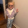 casual_clothing, child, cute, fashion, girl, hair_tied, happy, heart_shaped, indoor, jacket, jeans, lavender, person, pose, smile, sneakers, standing, sunglasses, wall, wooden_floor