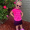 black_shorts, blonde_hair, brick_wall, casual_clothing, child, flowers, girl, green_leaves, hair_tie, happy, mulch, nature, outdoor, pink_shirt, pink_shoes, posing, rose_bush, smile, sunglasses, sunny