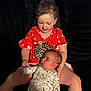 toddler, baby, child, sibling, holding, red_shirt, polka_dot, onesie, dinosaur_pattern, curly_hair, seated, black_background, indoor, candid, family, cute, affection, portrait, soft_lighting, young