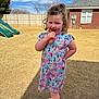 child, curly_hair, dress, colorful_clothing, outdoor, sunny, popsicle, happy, playful, smiling, grass, fence, house, window, sneakers, footwear, daylight, person, yard, playground