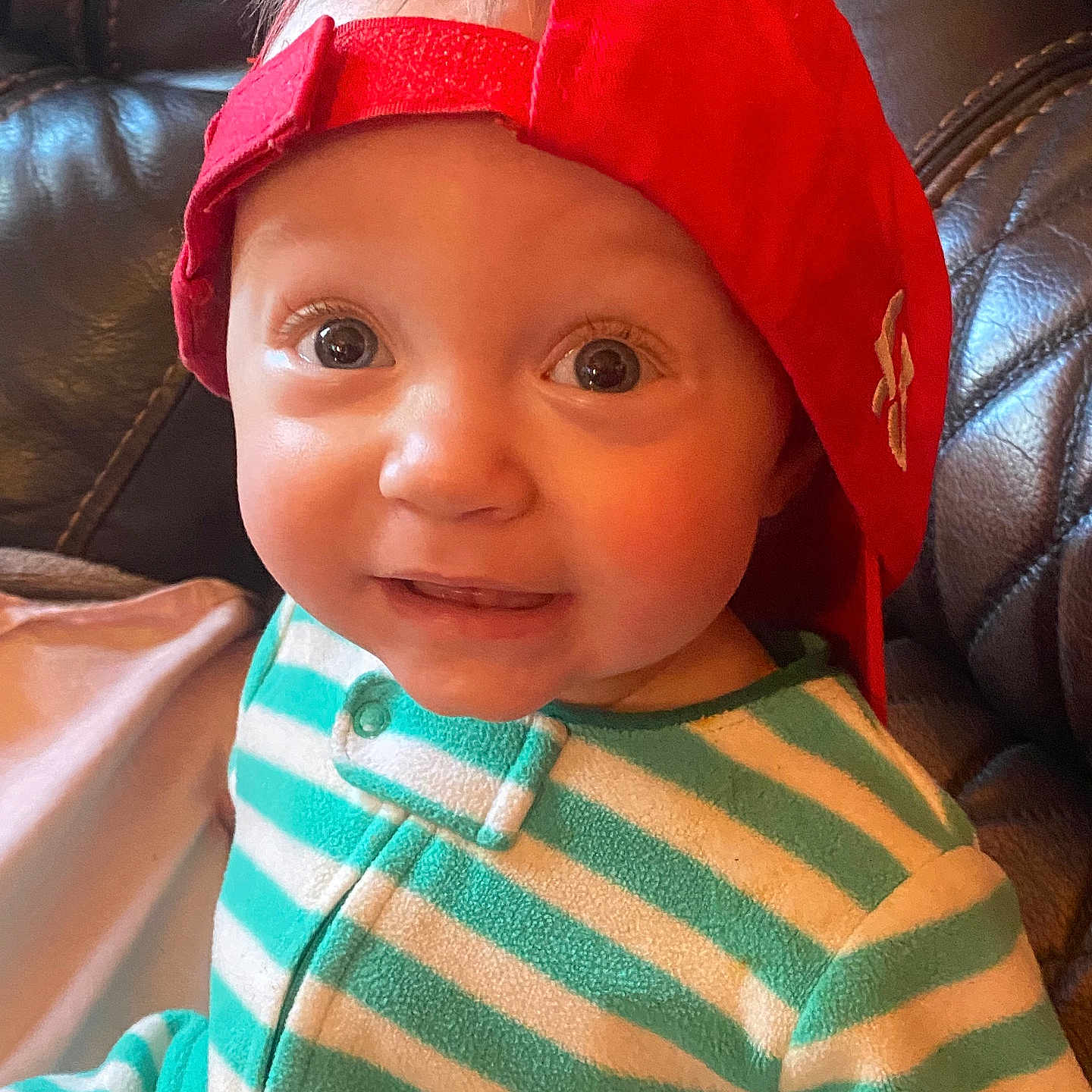 Trinity joined the competition — help win amazing prizes! baby, child, face, red_cap, striped_clothing, onesie, smile, curious, leather_couch, indoor, portrait, young_child, cute, headwear, person, infant, seated, happy, closeup, casual