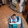 baby, bib, box, chair, child, cleaning_tools, clothing, curious, door, floor, furniture, home, indoor, living_room, person, smiling, table, tile_floor, toy, walker