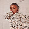 baby, child, infant, onesie, animal_print, hand_in_mouth, curly_hair, lying_down, white_background, quilted_bed, sleepwear, cute, portrait, indoors, small_child, face, expression, soft_lighting, comfortable, young