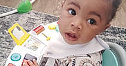 Amani is registered to the contest to win money with this photo: baby, child, infant, walker, toy, indoor, curious, bib, clothing, animal_print, face, hands, floor, rug, furniture, seat, expression, play, colorful, young