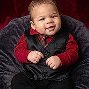 Michael is registered to the contest to win money with this photo: baby, infant, child, portrait, smiling, seated, formalwear, vest, tie, red_shirt, black_pants, chair, plush_fabric, studio, backdrop, closeup, fashion, adorable, hands, indoor