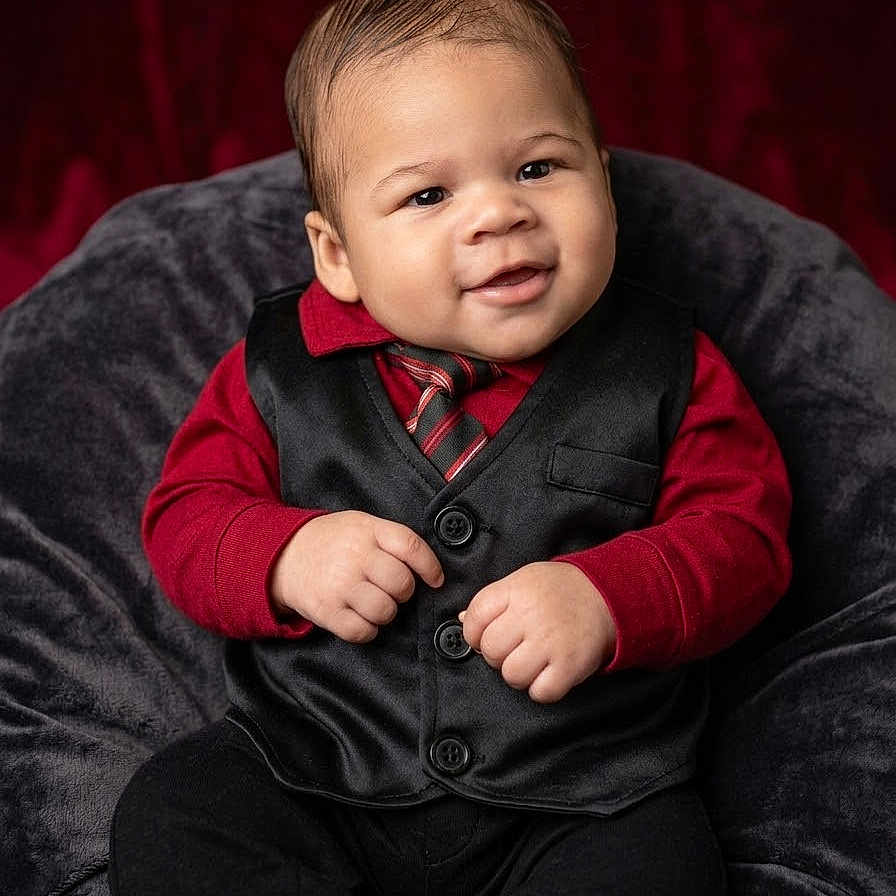 Michael is registered to the contest to win money with this photo: baby, infant, child, portrait, smiling, seated, formalwear, vest, tie, red_shirt, black_pants, chair, plush_fabric, studio, backdrop, closeup, fashion, adorable, hands, indoor