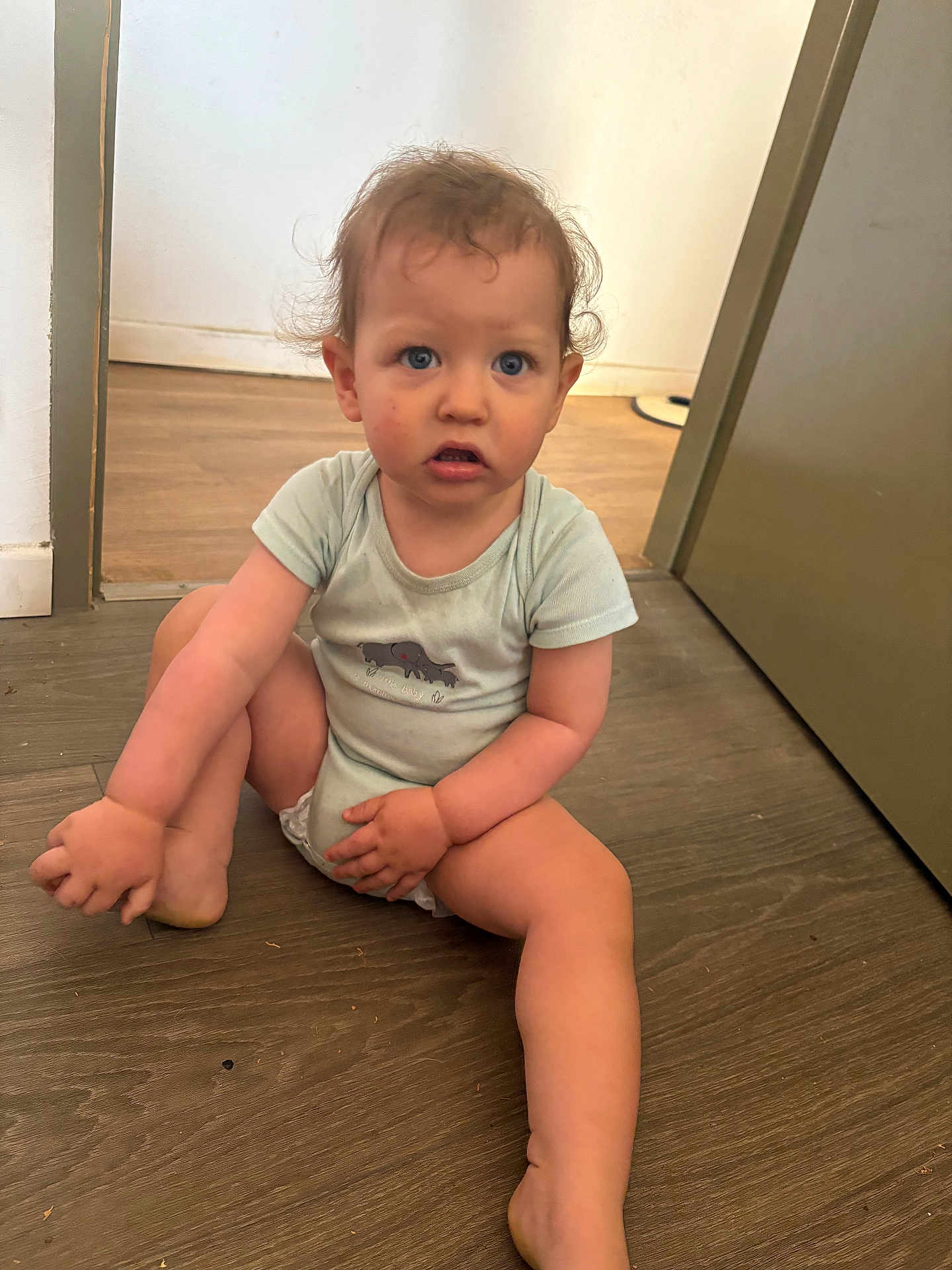 Lowen a rejoint le concours — aidez-le/la à gagner de superbes lots ! toddler, child, baby, sitting, indoors, wooden_floor, onesie, blue_eyes, curly_hair, holding_foot, door, wall, casual_clothing, expression, person, floor, young_child, cute, skin, hair