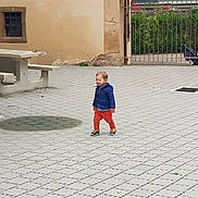 Lowen participe au concours pour gagner de l'argent avec cette photo : toddler, child, blue_jacket, orange_pants, walking, outdoor, courtyard, paved_ground, metal_gate, rustic_wall, picnic_table, greenery, daylight, casual_clothing, small_child, urban, sidewalk, person, footwear, young_child