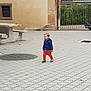 toddler, child, blue_jacket, orange_pants, walking, outdoor, courtyard, paved_ground, metal_gate, rustic_wall, picnic_table, greenery, daylight, casual_clothing, small_child, urban, sidewalk, person, footwear, young_child