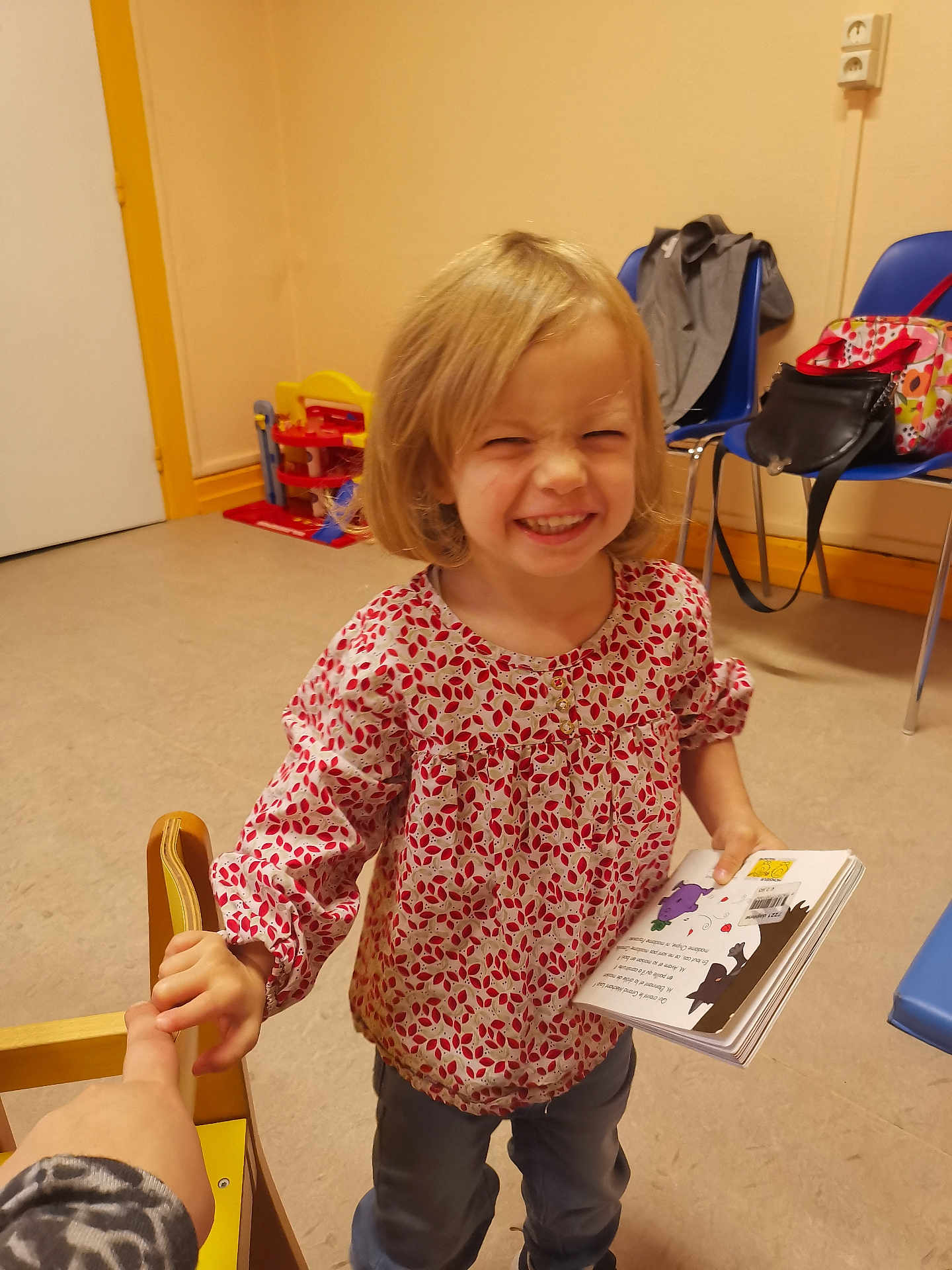 Santiana participe au concours pour gagner de l'argent avec cette photo : child, girl, smiling, holding_hand, book, indoor, toy, chair, playroom, casual_clothing, happy, person, blonde_hair, floor, wall, backpack, furniture, sleeves, young, cute
