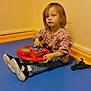 Santiana a rejoint le concours — aidez-le/la à gagner de superbes lots ! child, toy, indoor, blue_mat, red_toy, steering_wheel_toy, dinosaur_toy, casual_clothing, jeans, sneakers, light_hair, curious_expression, floor, corner, playtime, young_child, sitting, person, plastic_toy, yellow_border