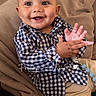 baby, child, smiling, clapping, checkered_shirt, beige_pants, couch, indoor, happy, cute, portrait, infant, clothing, hand_gesture, face, person, sitting, expression, home, cozy
