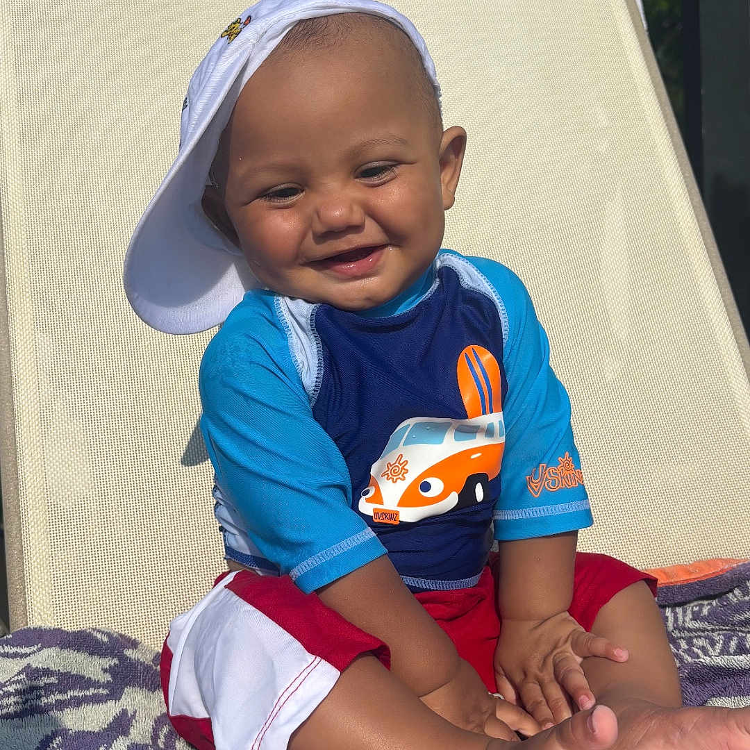 Kiel joined the competition — help win amazing prizes! toddler, child, smiling, hat, cap, swim_shirt, shorts, outdoor, sunlight, lounge_chair, towel, barefoot, happy, sitting, cute, summer, portrait, baby, clothing, relaxation