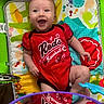 baby, infant, child, smiling, happy, portrait, lying_down, activity_center, playmat, toy, musical_toy, colorful, red_clothing, baseball_shirt, patterned_blanket, indoor, playroom, infant_seat, reflection, cheerful