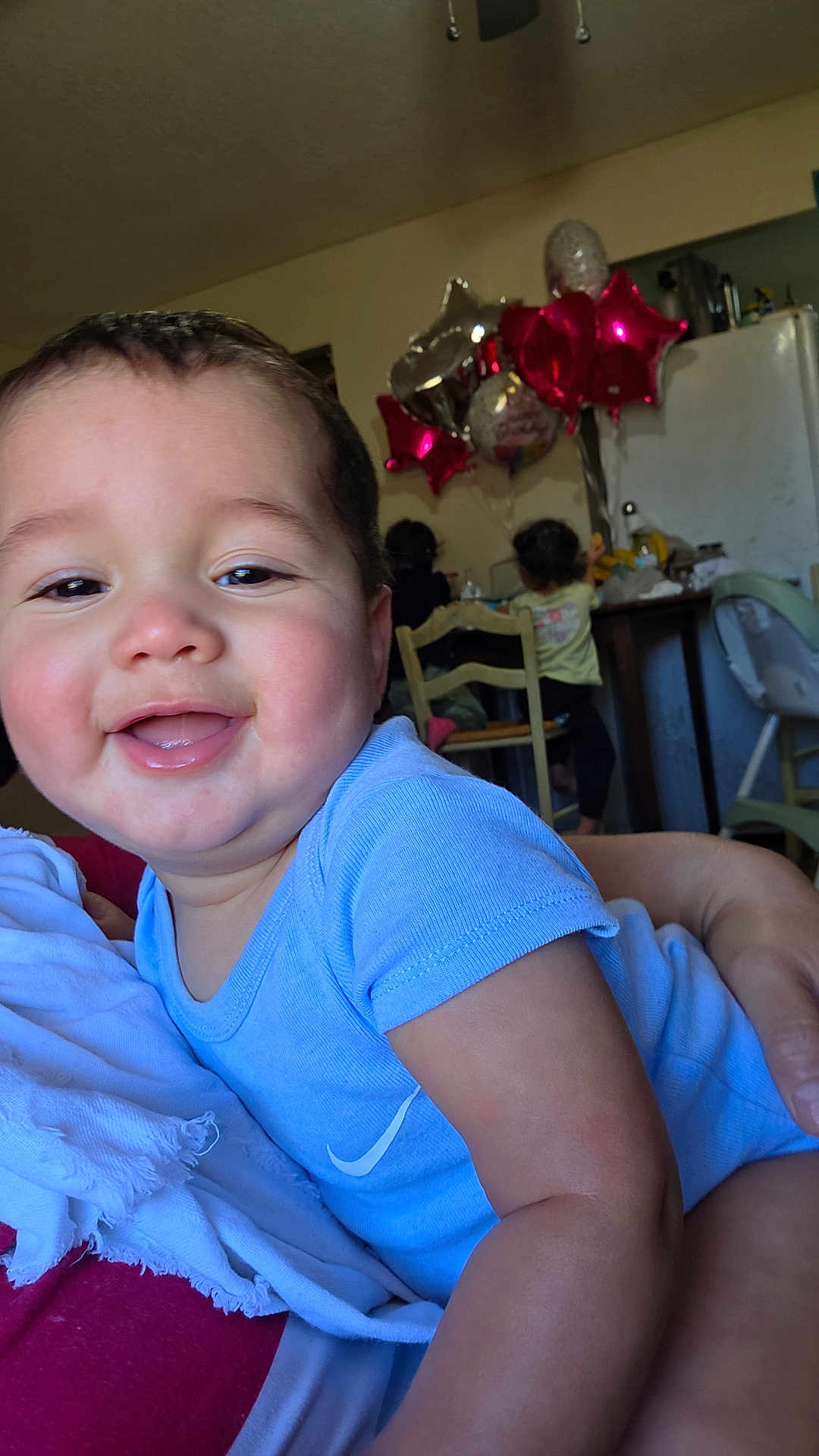 Asher Reyes joined the competition — help win amazing prizes! baby, child, toddler, balloons, blue_clothing, indoor, smiling, happy, person, face, chair, table, refrigerator, kitchen, celebration, family, holding, red_balloons, silver_balloons, home