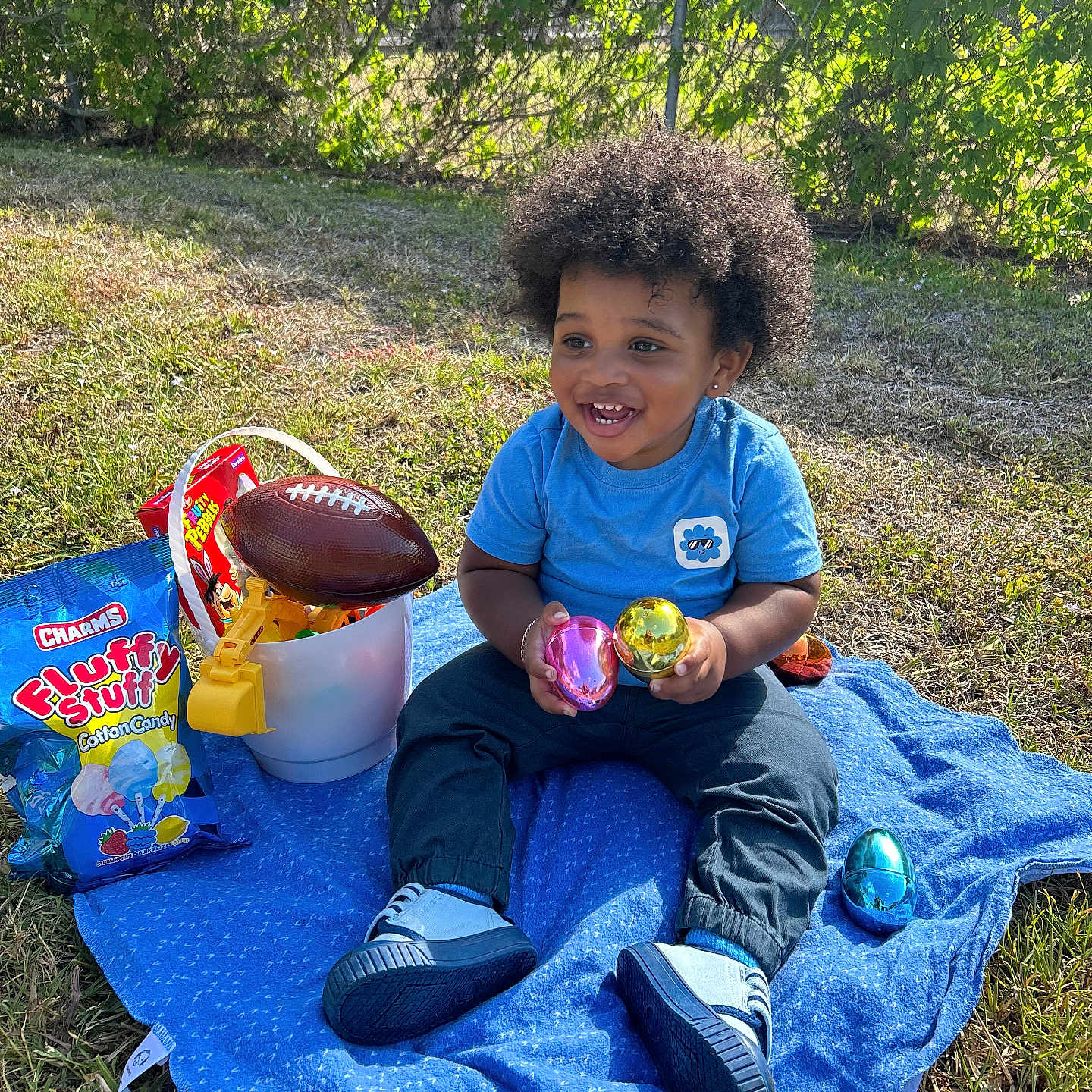 Khyri is registered to the contest to win money with this photo: afro_hair, blue_blanket, candy, casual_clothing, child, colorful_eggs, cute, football, fun, grass, happy, nature, outdoor, playtime, shoes, sitting, smiling, sunlight, toddler, toy_bucket