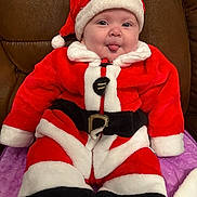 Killian is registered to the contest to win money with this photo: baby, child, santa_claus_costume, red_clothing, white_fur_trim, black_belt, hat, tongue_out, cute, smiling, indoor, couch, purple_blanket, plush, holiday, festive, portrait, person, seasonal, happy