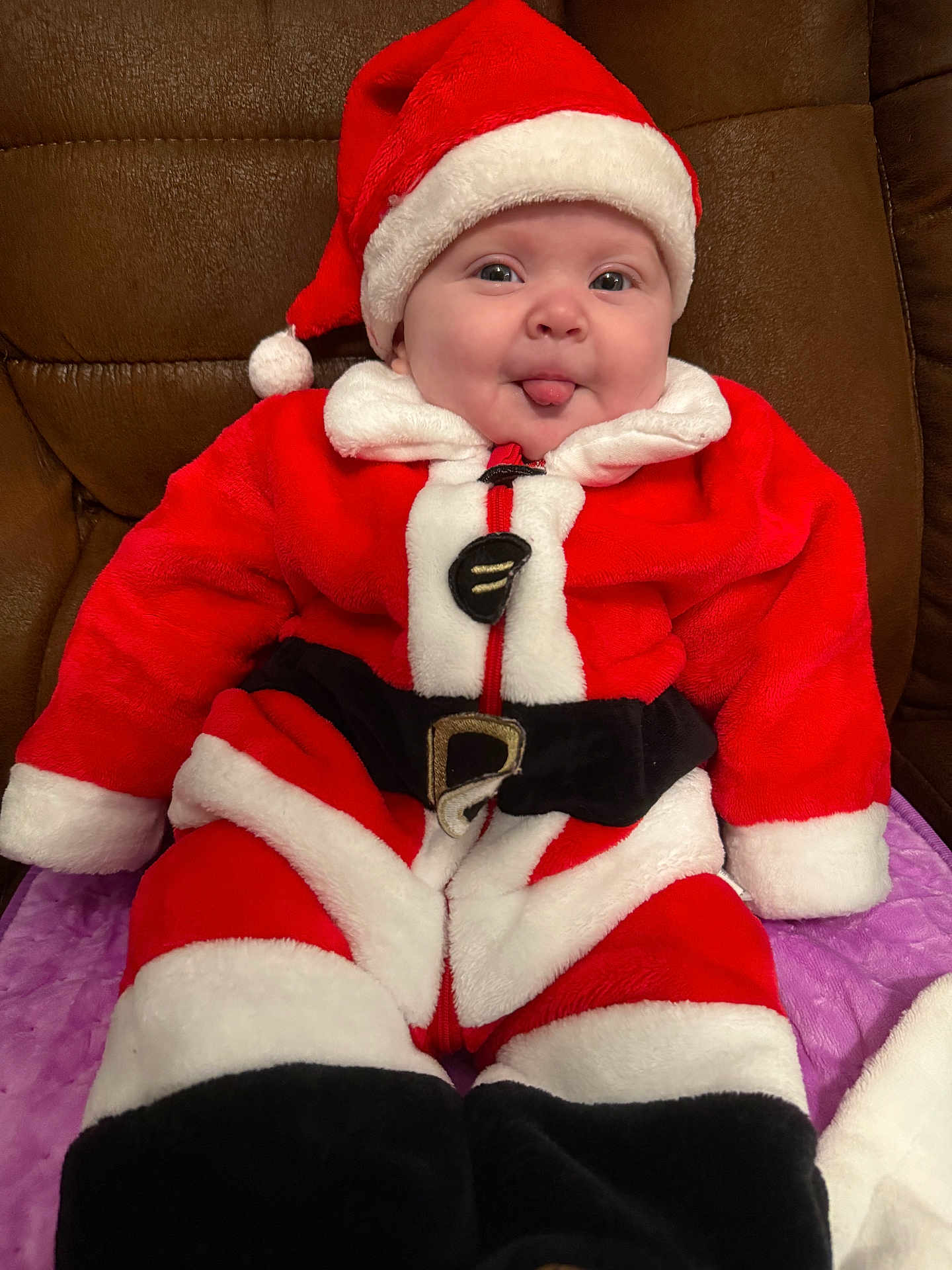 Killian is registered to the contest to win money with this photo: baby, child, santa_claus_costume, red_clothing, white_fur_trim, black_belt, hat, tongue_out, cute, smiling, indoor, couch, purple_blanket, plush, holiday, festive, portrait, person, seasonal, happy
