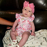 Harper joined the competition — help win amazing prizes! baby, infant, child, pink_outfit, headband, bow, blanket, leather_couch, footwear, sitting, person, indoor, cute, small_shoes, hand, support, patterned_blanket, expression, clothing, portrait