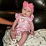 baby, infant, child, pink_outfit, headband, bow, blanket, leather_couch, footwear, sitting, person, indoor, cute, small_shoes, hand, support, patterned_blanket, expression, clothing, portrait