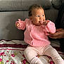 baby, child, pink_sweater, socks, curly_hair, smiling, bedspread, floral_pattern, hand, support, indoor, couch, seated, cute, portrait, warm_clothing, infant, cozy, person, home