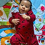 baby, infant, red_outfit, play_mat, colorful, cute, smiling, curly_hair, indoor, child, person, lying_down, patterned_clothing, bright_colors, soft_toys, background, happy, young_child, sock, playtime