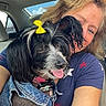 woman, dog, denim_jacket, yellow_bow, car_interior, happy, smiling, pet, portrait, close_up, casual_clothing, seat, window, sunlight, tongue_out, hair, face, blue_shirt, star_pattern, affection