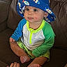 baby, child, hat, bucket_hat, fish_pattern, tongue_out, sitting, couch, brown_couch, colorful_clothing, long_sleeve_shirt, green_sleeves, shorts, barefoot, hand, indoor, cute, playful, face, person
