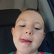 Daniel is registered to the contest to win money with this photo: child, face, tongue, green_shirt, car_seat, short_hair, playful, cheeky, portrait, person, indoors, seat, clothing, young, expression, head, skin, casual, childhood, vehicle