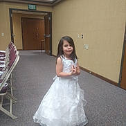 Melony joined the competition — help win amazing prizes! child, girl, white_dress, carpet, chairs, indoor, room, door, wall, exit_sign, long_hair, standing, smiling, young, person, floor, light, furniture, empty_room, wall_socket