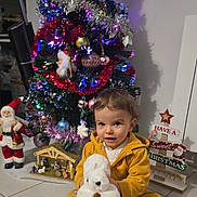 Gina a rejoint le concours — aidez-le/la à gagner de superbes lots ! toddler, child, yellow_clothing, stuffed_animal, white_plush, christmas_tree, holiday_decorations, santa_figure, nativity_scene, floor_tiles, holiday_spirit, indoors, festive_lights, ornaments, smiling_child, seasonal, celebration, cute, home, christmas_sign