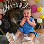 Elissa is registered to the contest to win money with this photo: baby, child, blue_dress, balloons, party, celebration, snack, smiling, person, footwear, frilly_shoes, happy, indoor, people, headband, number_balloon, pink_balloons, yellow_balloons, white_balloons, festive