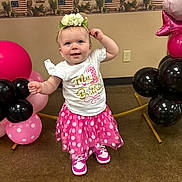 Adalynn is registered to the contest to win money with this photo: baby, child, birthday, first_birthday, pink, white, shirt, skirt, sneakers, flower_crown, balloons, polka_dot, celebration, smiling, standing, indoor, floor, wall, decor, face