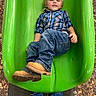 child, boy, slide, playground, green_slide, jeans, boots, plaid_shirt, outdoor, fall_leaves, smile, casual_clothing, fun, recreation, daylight, person, play, clothing, nature, happy