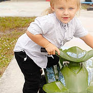 Wyatt is registered to the contest to win money with this photo: child, boy, toy_vehicle, outdoor, sidewalk, grass, casual_clothing, short_sleeves, sneakers, blond_hair, blue_eyes, playful, curious_expression, standing, holding, plastic_toy, daylight, background_blur, concrete, young