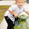 child, boy, toy_vehicle, outdoor, sidewalk, grass, casual_clothing, short_sleeves, sneakers, blond_hair, blue_eyes, playful, curious_expression, standing, holding, plastic_toy, daylight, background_blur, concrete, young