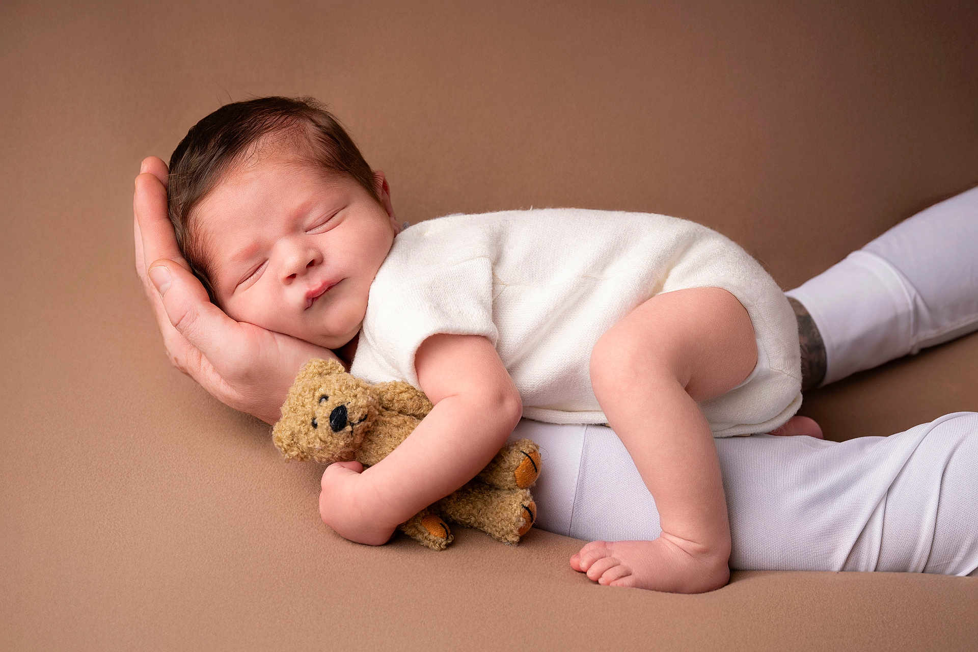 Léo participe au concours pour gagner de l'argent avec cette photo : baby, newborn, sleeping_baby, teddy_bear, hand, parent_hand, white_clothing, brown_background, portrait, studio, soft_texture, cozy, infant, cute, peaceful, bare_foot, toes, face, head, plush_toy