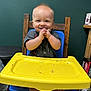 baby, blue_chair, child, clapping_hands, face, food, foot, furniture, green_wall, happy, high_chair, home, indoor, messy, person, shirt, smiling, table, wooden_chair, yellow_tray