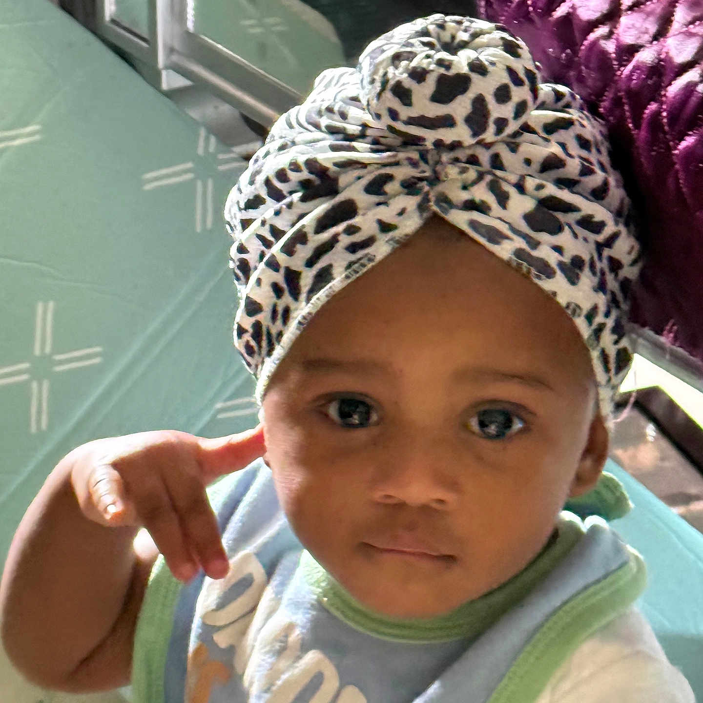 Sianni is registered to the contest to win money with this photo: baby, infant, child, portrait, face, eyes, serious_expression, headwrap, leopard_print, bow, bib, shirt, sitting, indoor, bedroom, furniture, quilt, sunlight, close_up, photography