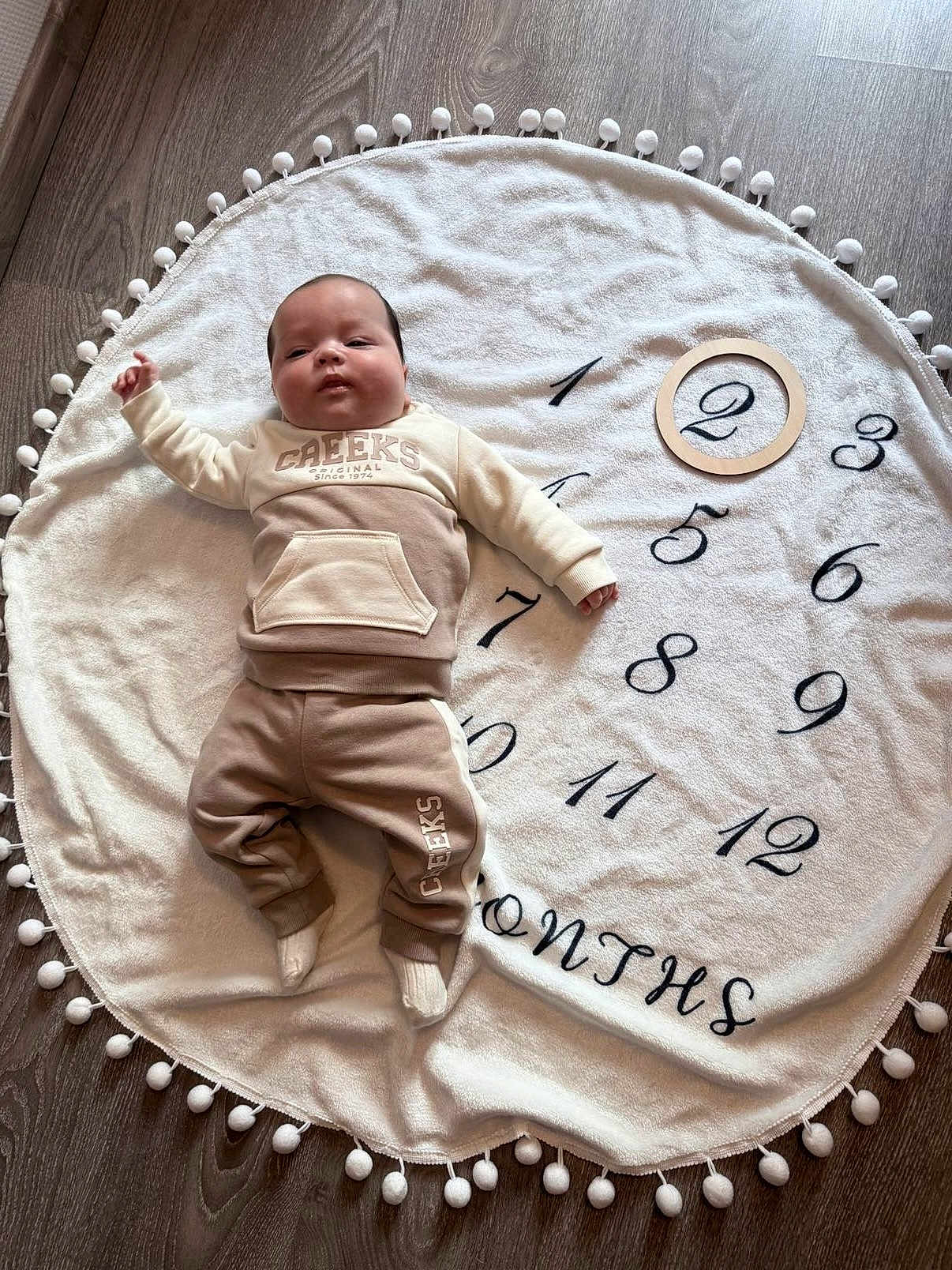Jules a rejoint le concours — aidez-le/la à gagner de superbes lots ! baby, infant, blanket, numbers, months, wood_floor, clothing, socks, face, person, lying_down, round_blanket, wooden_ring, flooring, indoor, cozy, cute, child, portrait, age_marker