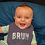 baby, smiling, bib, text_on_bib, blue_cushion, green_outfit, short_sleeves, happy, child, person, indoors, face, head, mouth_open, light_skin, small_hands, seated, clothing, infant, expression